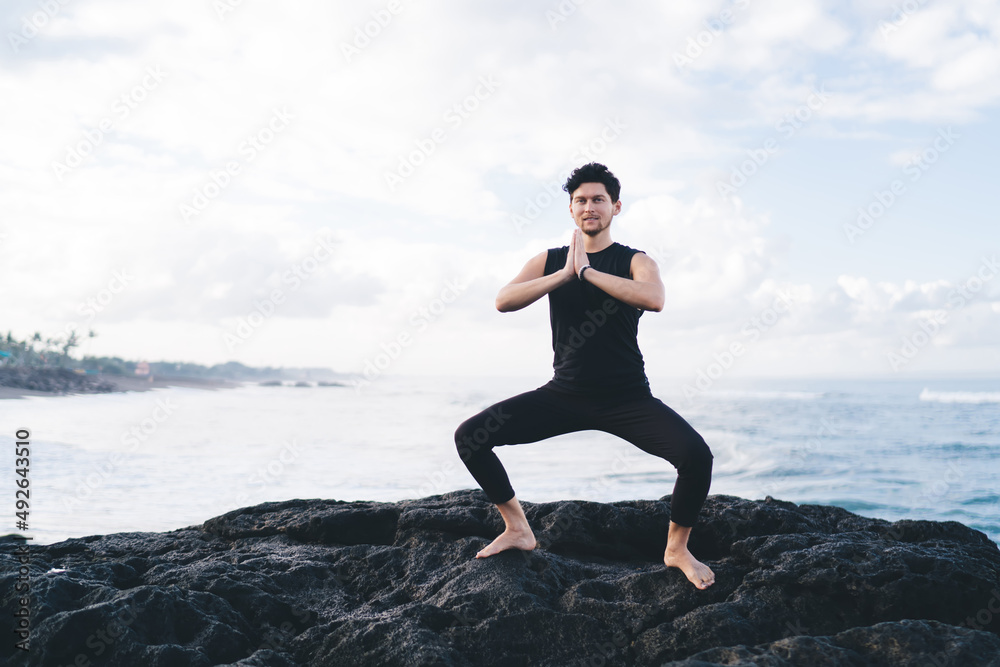Caucasian man in black sportswear looking at camera during mediation time and hatha yoga practice, portrait of healthy male enjoying leisure for namaste enlightenment at coastline seashore