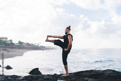 Side view of pumped up male yogi recreating doing balance yoga poses feeling inner peace and vitality, male with strong body standing in asana care about physical and mental health at seashore