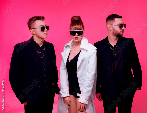 Front view of female performer with red lips and dark sunglasses, dressed in black underwear and white long trench coat, standing between two men and looking at camera.  photo