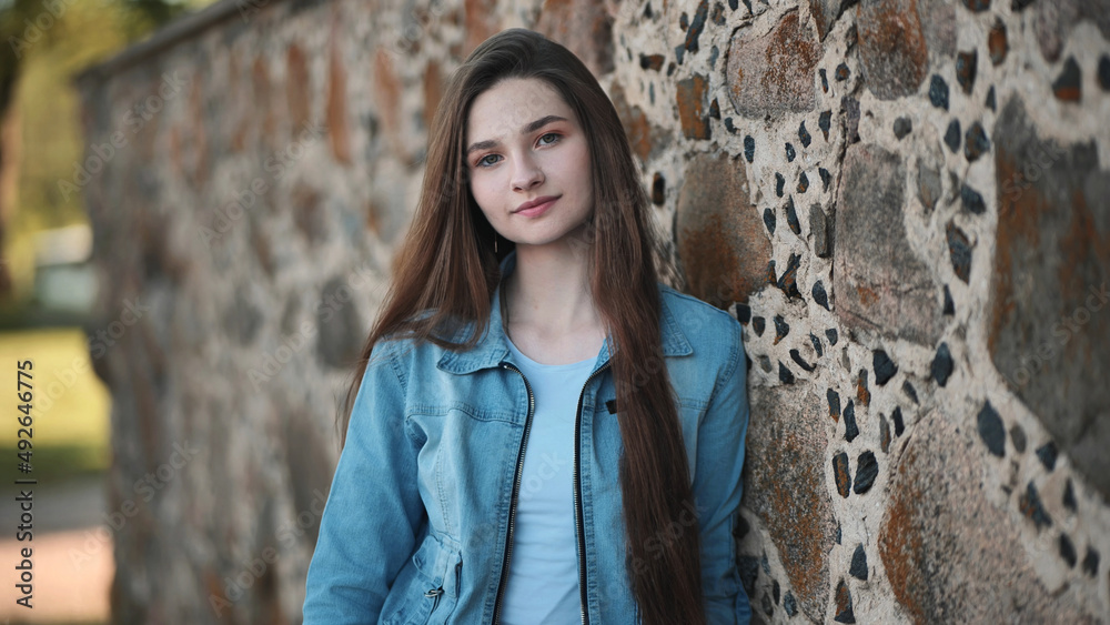 Portrait of a long-haired girl in a denim jacket against a stone wall.