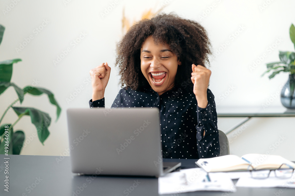 Surprised successful confident emotional african american business woman, broker, manager, sitting at table with laptop, enjoying big profit, victory, good news, gesturing with fists, smiling Surprised successful confident emotional african american business woman, broker, manager, sitting at table with laptop, enjoying big profit, victory, good news, gesturing with fists, smiling