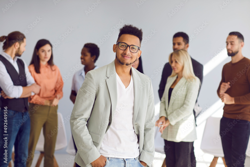 Young black man in suit jacket, casual tee and stylish glasses standing hands in pockets in front of diverse coworkers. Portrait of handsome businessman, team leader or business conference participant Young black man in suit jacket, casual tee and stylish glasses standing hands in pockets in front of diverse coworkers. Portrait of handsome businessman, team leader or business conference participant
