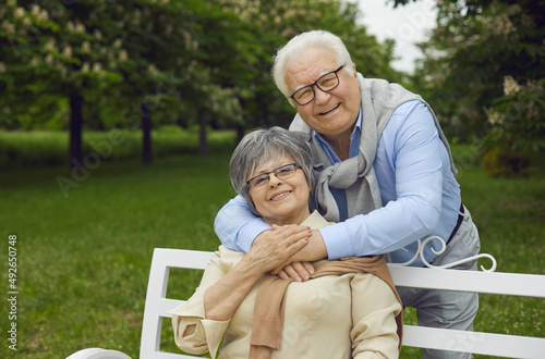 Elderly happy man and woman are resting in the park on a bench, breathing fresh air, a man standing behind and holding his spouse, happy family life, outdoor recreation. © Studio Romantic