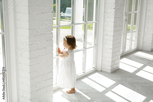 A little barefoot girl stands at a large panoramic window and waits for her parents to return. The child looks out the window alone. A clean, bright room with natural sunlight. 