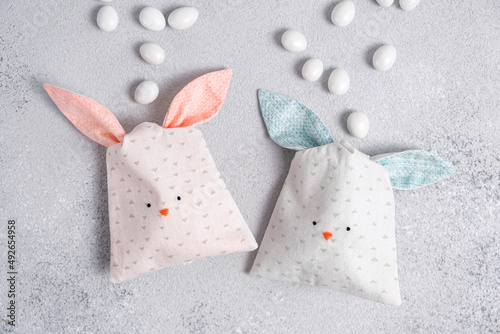 Two bag of sweets in shape of Easter bunny with blue and pink ears on light gray background. Packing of cotton fabric in form of cute bunnys or rabbits, sewing for children. Scattered chocolate eggs.