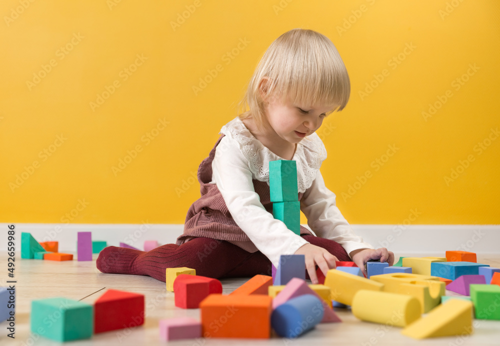 A little focused girl is sitting on the floor and building tower of multicolored blocks of the constructor. Selective focus A little focused girl is sitting on the floor and building tower of multicolored blocks of the constructor. Selective focus