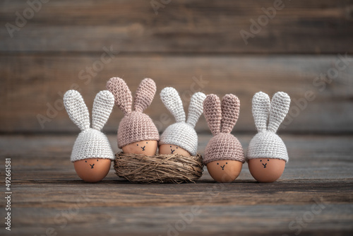 Five Easter eggs in crocheted hats with rabbit ears on a rustic wooden table. Soft focus.