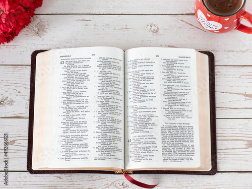 Open Holy Bible Book with a cup of coffee, and red flowers on a wooden table. Top view. Biblical concept of morning prayer, devotion, faith, and study from Scriptures inspired by God Jesus Christ. 