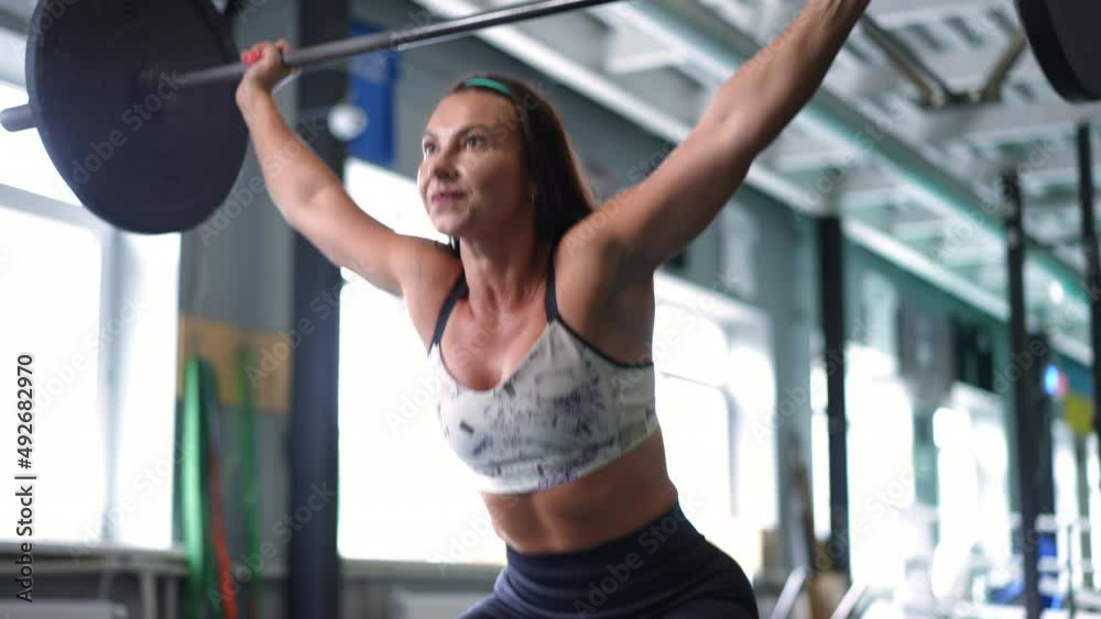 Smiling fit sportswoman squatting with barbell in gym in slow motion looking away. Portrait of confident athletic Caucasian woman training working out with heavy equipment indoors Smiling fit sportswoman squatting with barbell in gym in slow motion looking away. Portrait of confident athletic Caucasian woman training working out with heavy equipment indoors