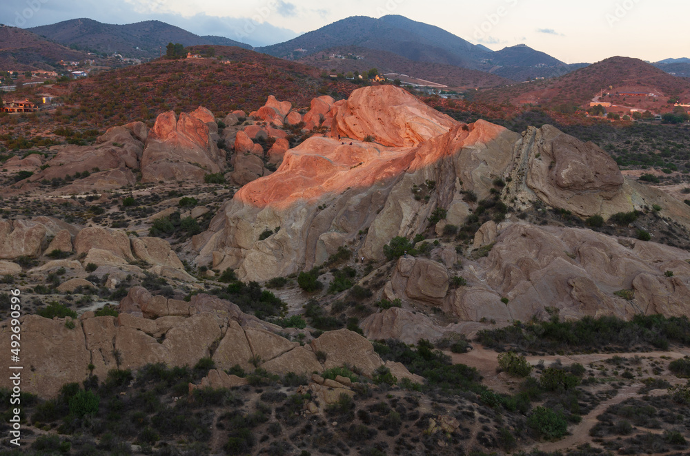 Vasquez Rocks Natural Area, taken from a nearby hill at the time of sunset. The sun rays selectively illuminate the middle ground area. A number of homes are shown in the background.