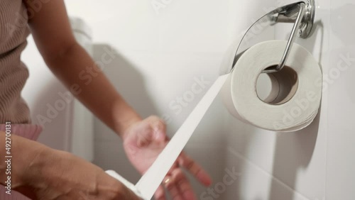 Closeup hand pulling toilet paper roll in holder for wipe, woman sitting on toilet she taking and tearing white tissue on wall to towel clean in bathroom, Healthcare concept