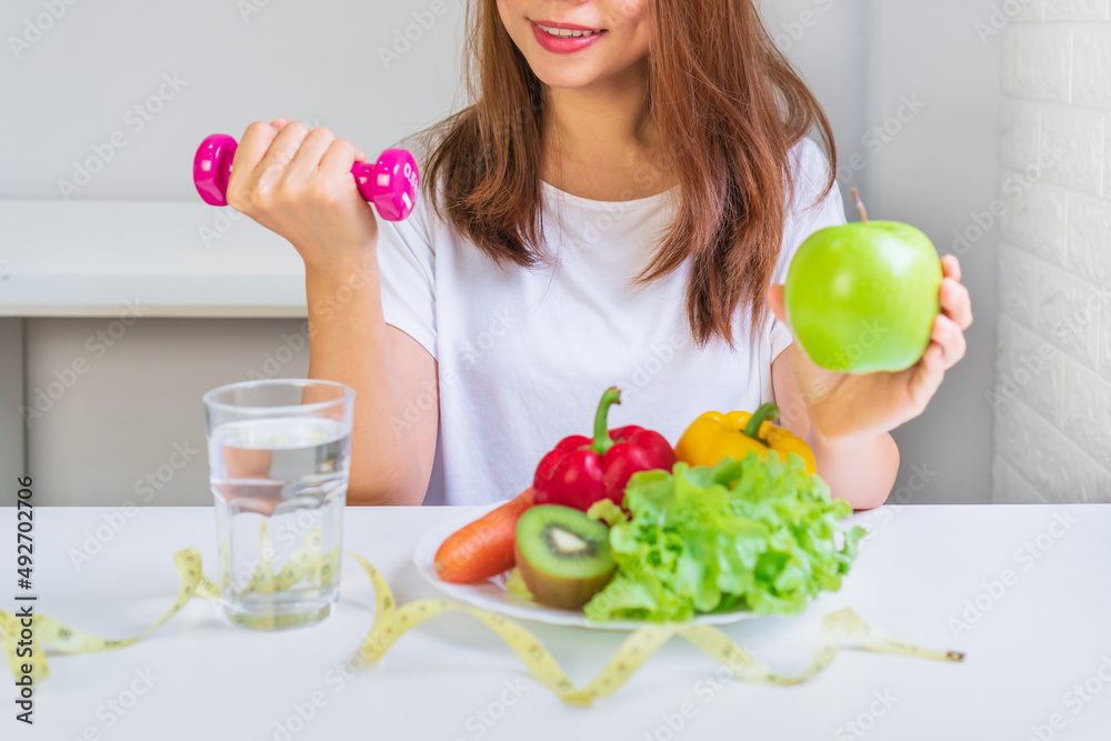 Women hands holding green apple while holing dumbbell for exercise with fruits, vegetables, water and tape measure on white table background. Selection of healthy food and exercise concept. Women hands holding green apple while holing dumbbell for exercise with fruits, vegetables, water and tape measure on white table background. Selection of healthy food and exercise concept.