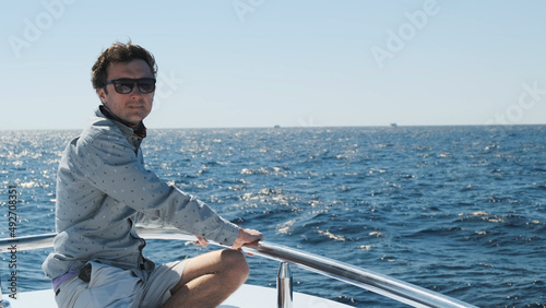 Man sits on yacht bow, looks into distance backlit morning sun rays. Yacht sailing on blue sea under clear sky. Successful handsome caucasian man in light clothes enjoys summer vacation on white yacht