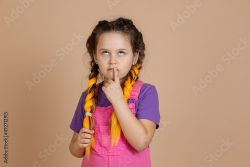 Portrait of pensive young female with yellow kanekalon braids, with eyes looking up touching mouth with finger wearing pink jumpsuit and purple t-shirt on beige background. photo