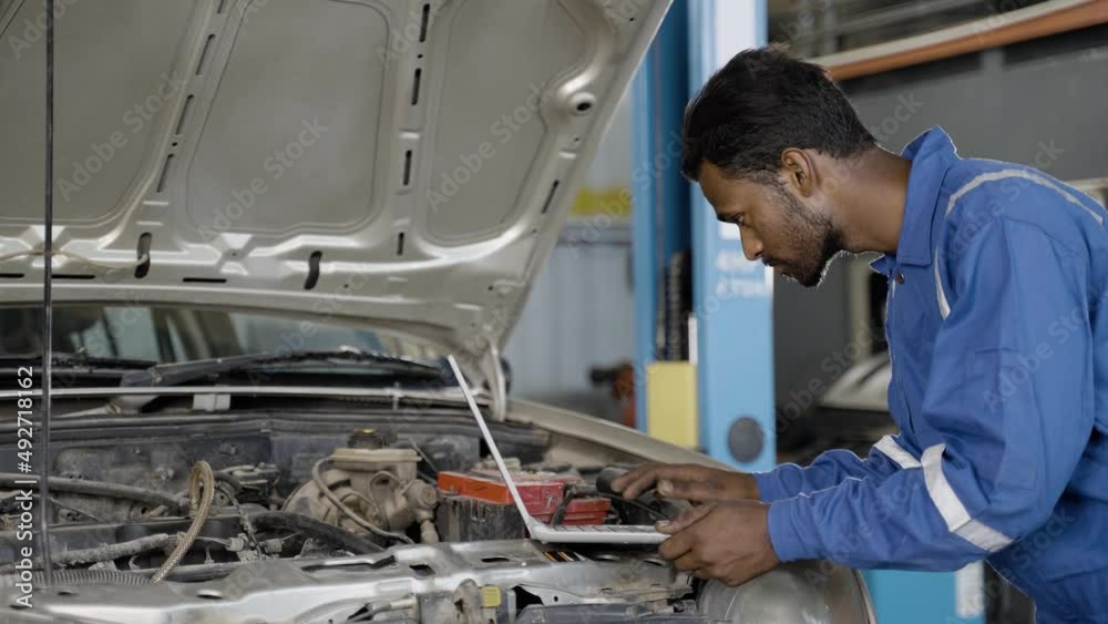 Car mechanic using laptop while checking car engine or circuit at garage - concept of professional, technology, expertise and maintance service.