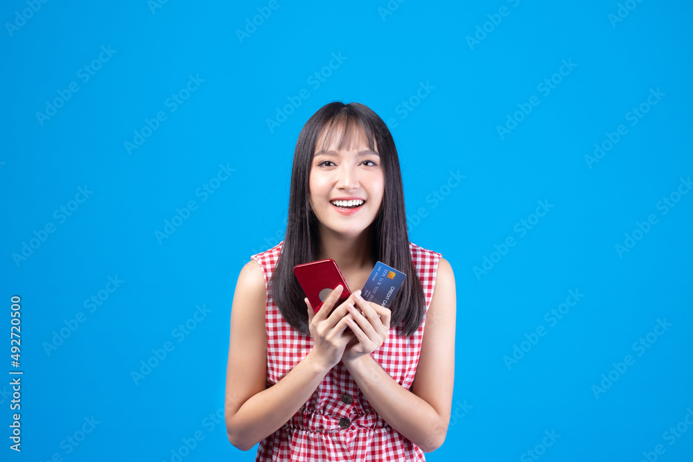 Portrait Asian happy young girl smiling cheerful and showing plastic credit card while holding mobile phone isolated on blue background Portrait Asian happy young girl smiling cheerful and showing plastic credit card while holding mobile phone isolated on blue background