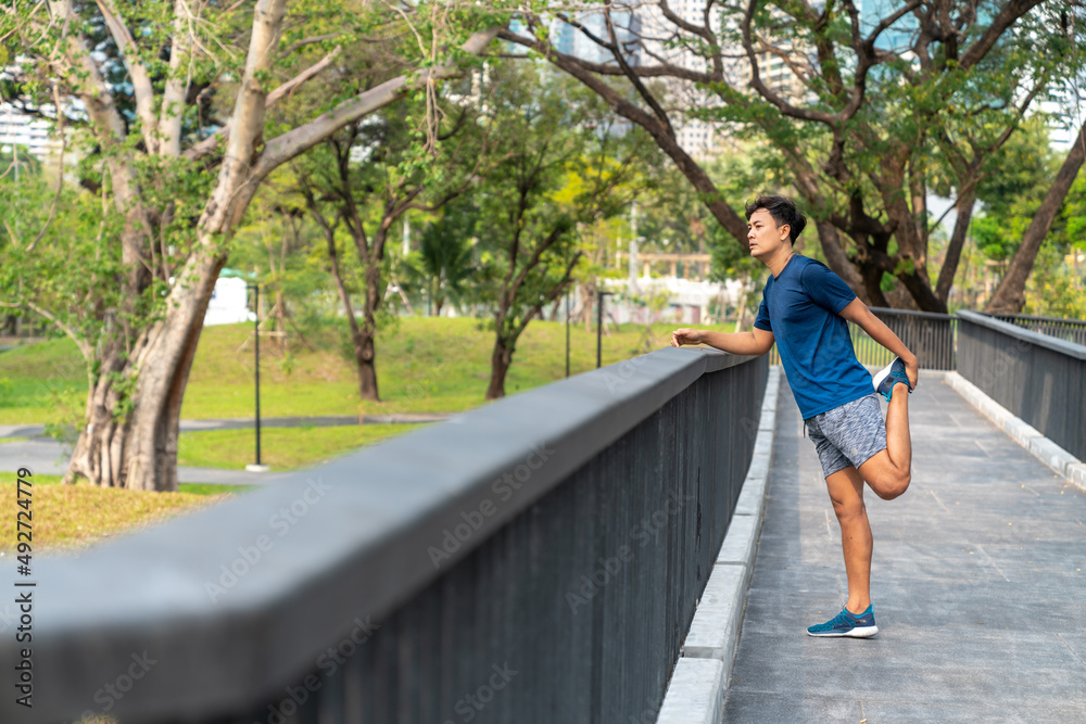 Asian man in sportswear and smartwatch stretching body while jogging at public park in the city in the morning. Healthy male athlete enjoy outdoor lifestyle sport training workout running exercise