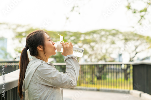 Young Asian woman in sportswear drinking water from a bottle while jogging at city public park in the morning. Healthy female athlete enjoy outdoor lifestyle sport training workout running exercise