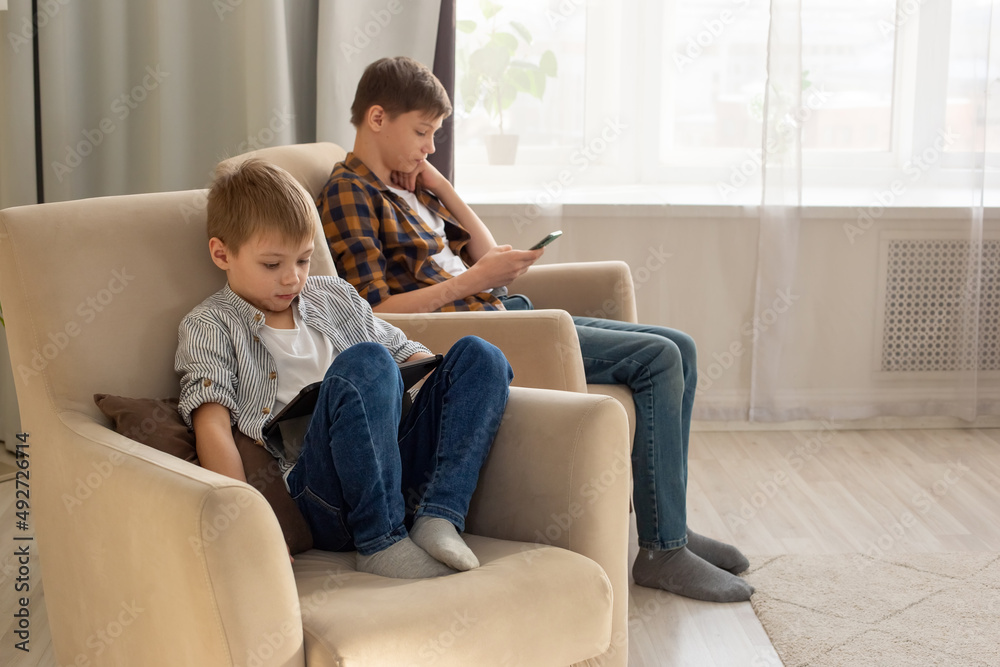Two boys, a child and a teenager, sit in beige armchairs, in a room, during the day, play their gadgets Two boys, a child and a teenager, sit in beige armchairs, in a room, during the day, play their gadgets