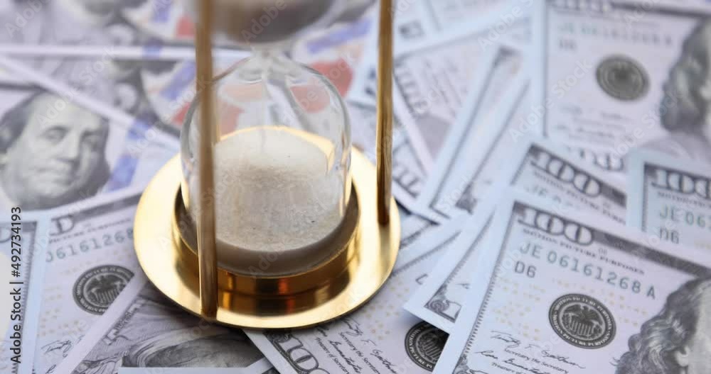 Sand timer clock with sparkling white sand and a person putting 100 dollar American banknotes. Concept: Saving money investing