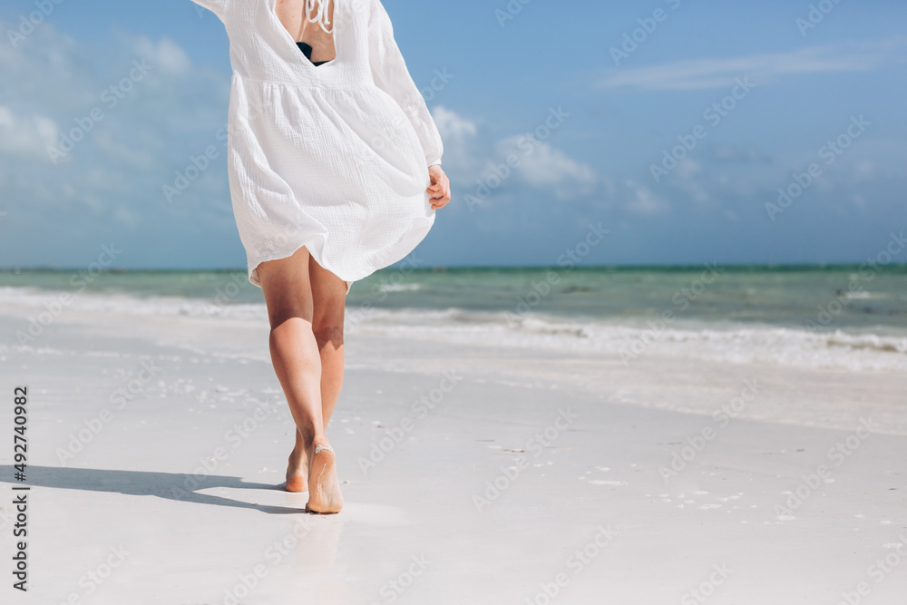 Elegant woman in white dress on luxury beach vacation relaxing in summer tropical ocean background. Relax feet and legs walking enjoying sun tan lifestyle.