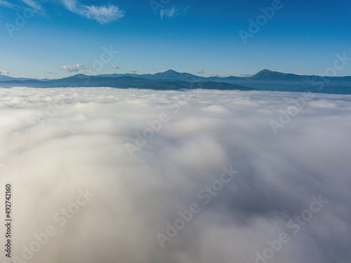 Flight over fog in Ukrainian Carpathians in summer. A thick layer of fog covers the mountains with a solid carpet. Mountains on the horizon. Aerial drone view. © Sergey