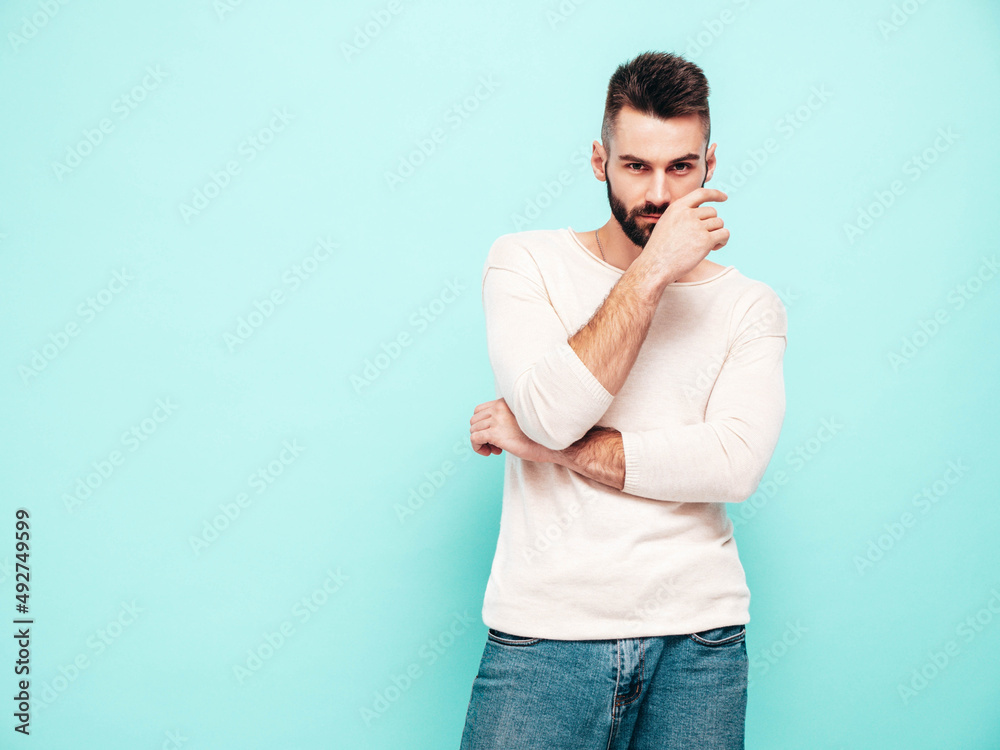 Portrait of handsome confident stylish hipster lambersexual model.Man dressed in white sweater and jeans. Fashion male isolated on blue wall in studio. Thoughtful Portrait of handsome confident stylish hipster lambersexual model.Man dressed in white sweater and jeans. Fashion male isolated on blue wall in studio. Thoughtful