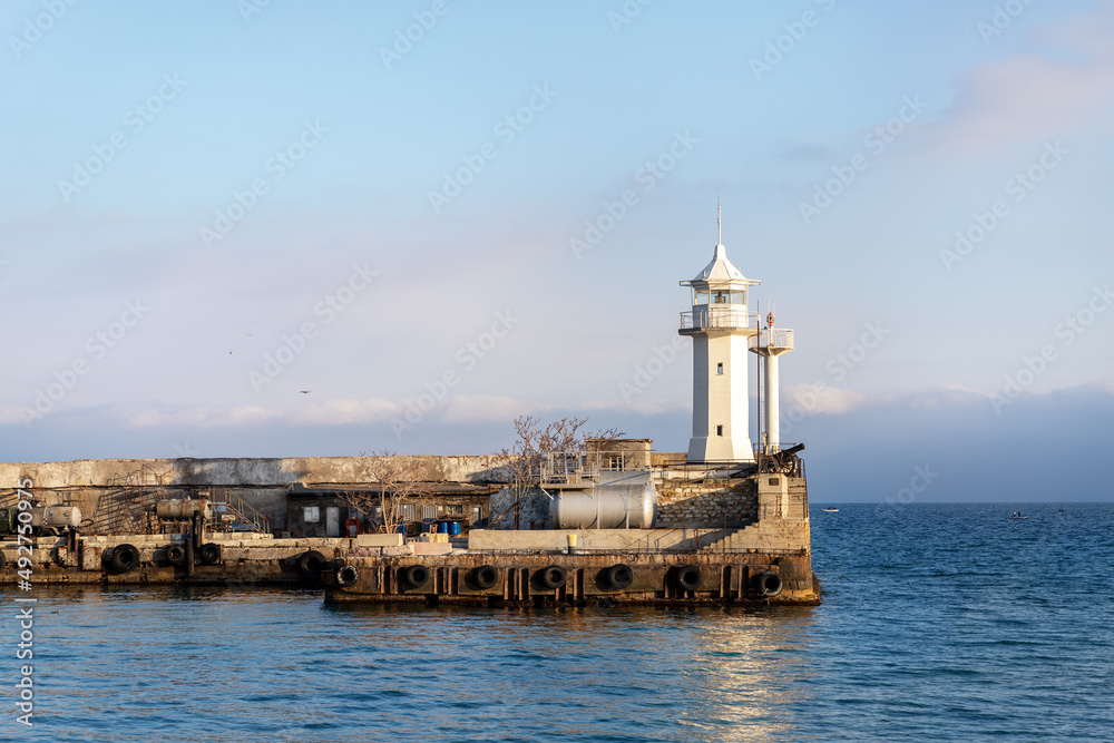 Scenic cityscape view old ancient white lighthouse building on stone pier at Yalta Crimean harbor on Black sea on warm sunny day. Bay nautical beacon house tower