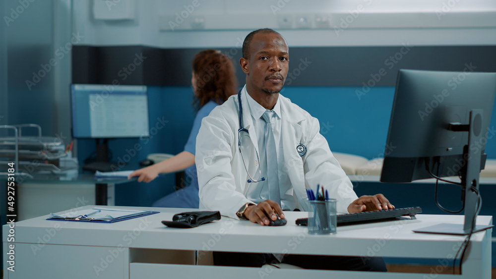 Obraz premium Portrait of health specialist working on computer at desk, wearing stethoscope and white coat in medical office. Male doctor using monitor to plan consultation and looking at camera.