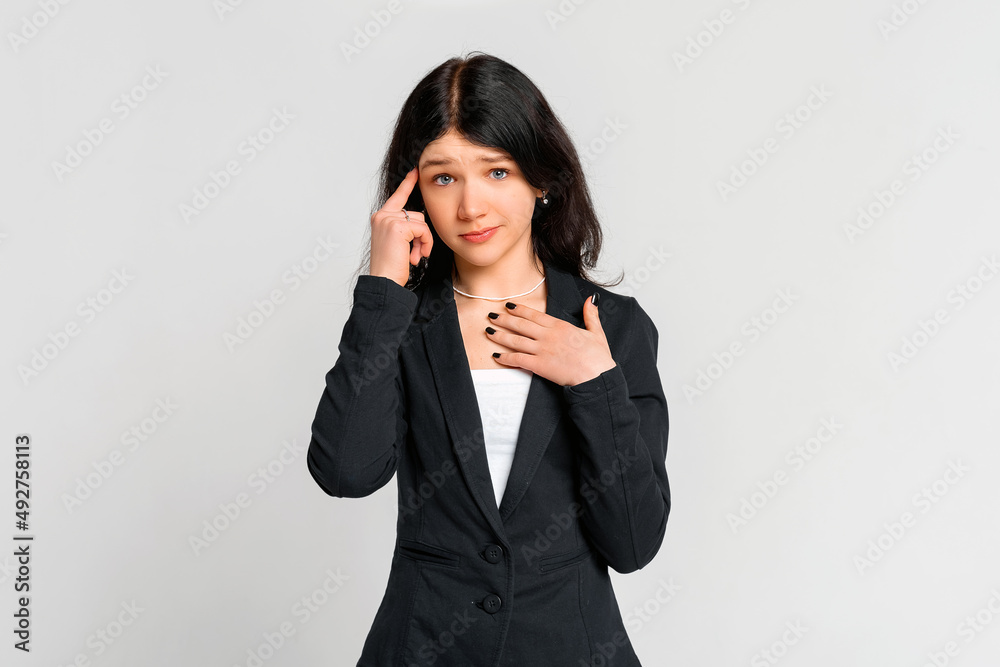 Embarrassed young dark-haired girl thinking, holding finger at her temple and looking insecure at camera, standing in black blazer over grey background Embarrassed young dark-haired girl thinking, holding finger at her temple and looking insecure at camera, standing in black blazer over grey background