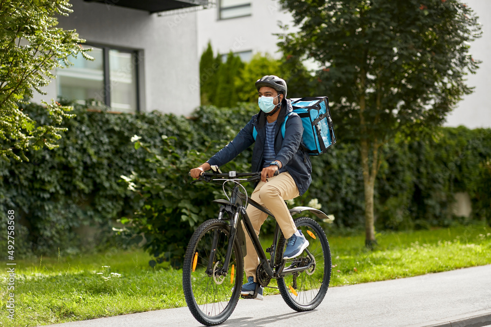 food shipping, health and people concept - delivery man in bike helmet and protective medical mask with thermal insulated bag and smatphone riding bicycle on city street