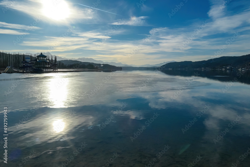 Fototapeta premium View on Woerthersee in Poertschach in Carinthia, Austria.The calm surface of the lake is reflecting the mountains, sunbeams and clouds. Clear and sunny day. Karawanks mountain range. Alps, Lake Woerth