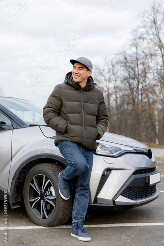 A young cheerful guy in a jacket and a cap stands near a new car photo