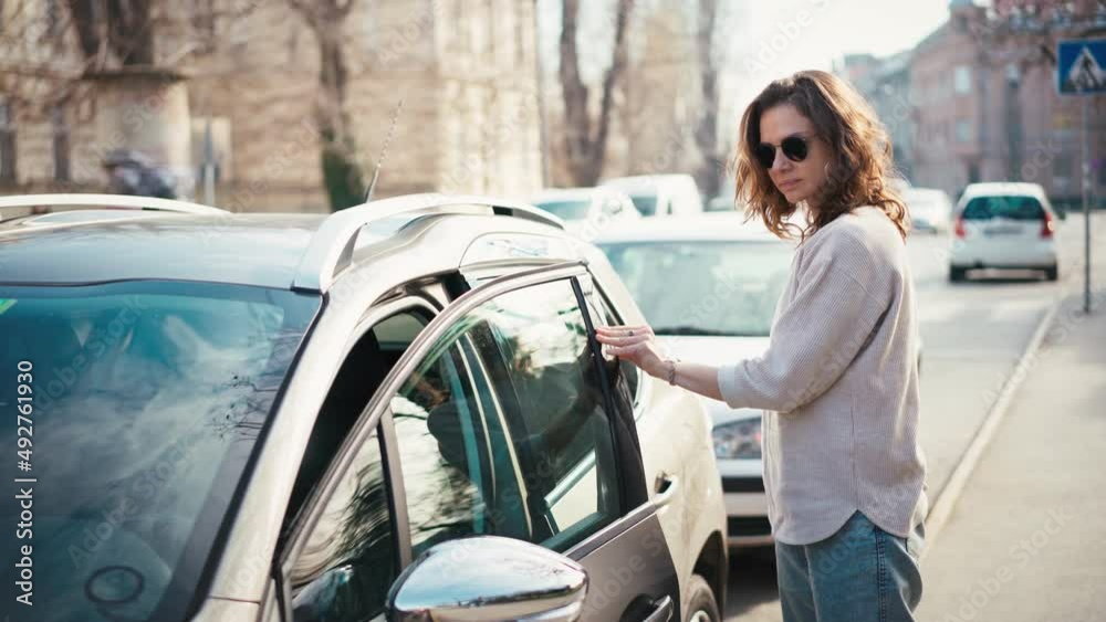 A young woman in sunglasses gets out of a parked car and locks it with a remote key.