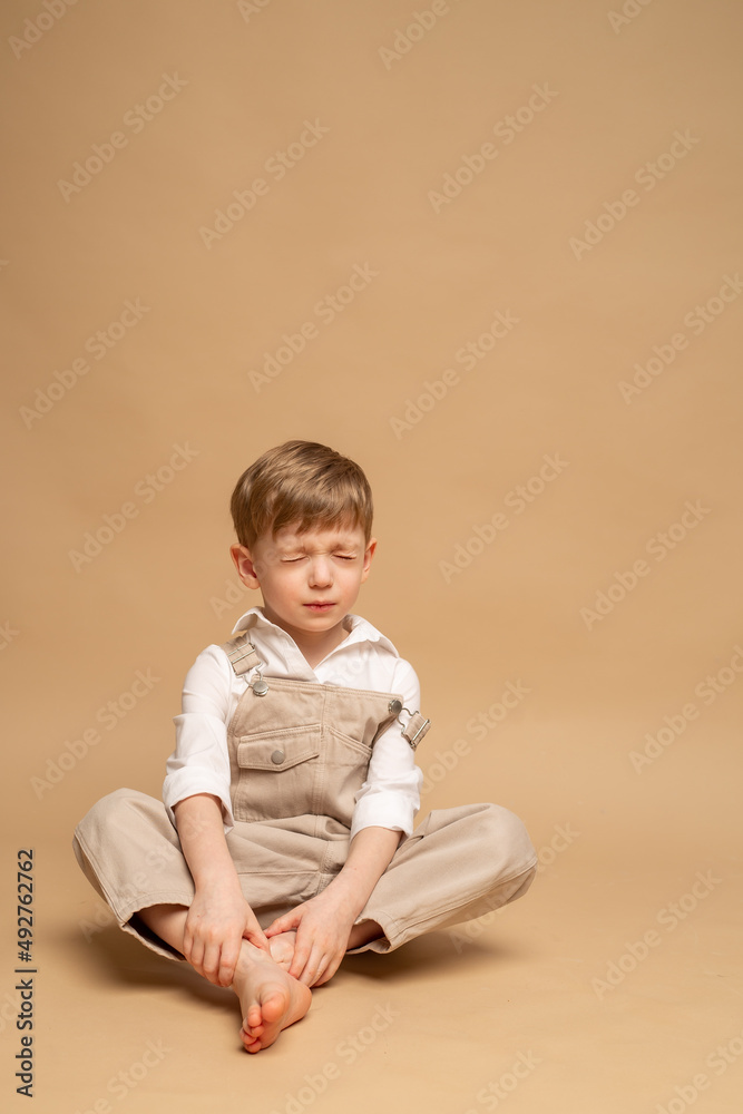 a charming blond four-year-old boy in beige overalls and a white shirt sits on a beige background a charming blond four-year-old boy in beige overalls and a white shirt sits on a beige background
