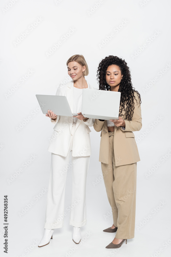 Full length of stylish interracial businesswomen using laptops on grey background. Full length of stylish interracial businesswomen using laptops on grey background.