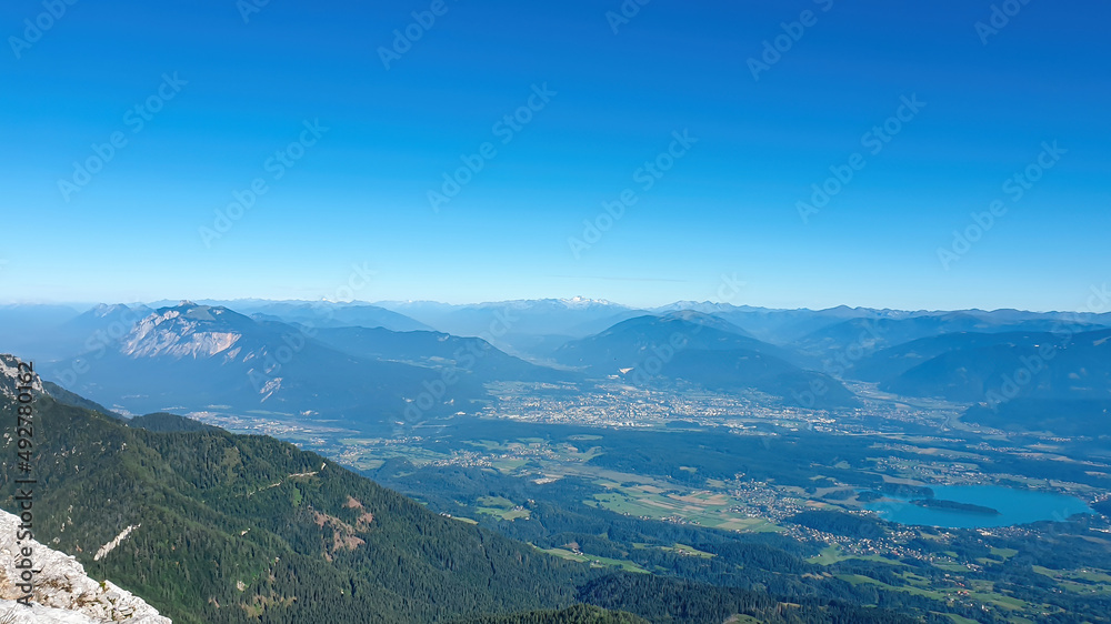 A panoramic view on the Alps from the top of Mittagskogel in Austrian Alps. Clear and sunny day. There is a lake at the bottom. A bit of haze in the valley. Outdoor activity. Alpine mountain chains