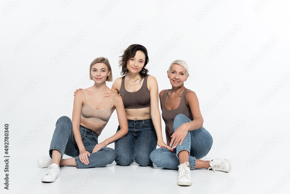 Women in tops and jeans looking at camera on grey background.