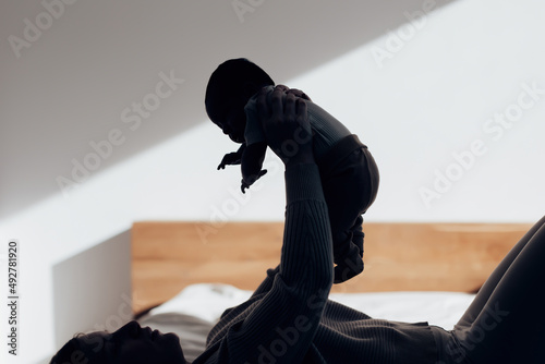 backlit silhouette of a mum lying in bed holding newborn baby in her hands lifting him up - natural daylight motherhood concept
