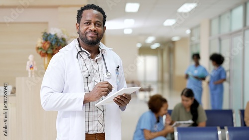 Portrait of black curly haired doctor with white gown uniform holding medical record smiling to camera standing in examination room working in a physician. Black people physician at hospital.