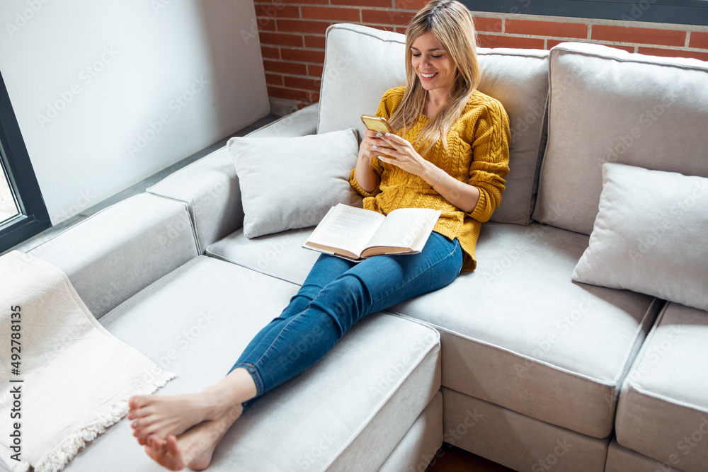Obraz premium Pretty young woman reading a book while sitting on sofa at home.