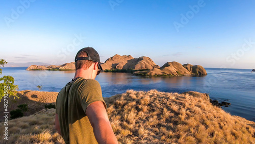 A man walking on top of a small island, enjoying the morning sun over Komodo National Park, Flores, Indonesia. Golden hour over the islands and sea. He is holding a selfie stick and taking pictures.