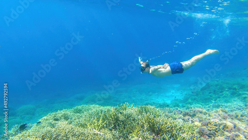 A man in masker and fins snorkelling in a vivid coral reef in Komodo National Park, Indonesia. The man is diving to see the reef from closer. Crystal clear water. Air bubbles around him. Free diving.