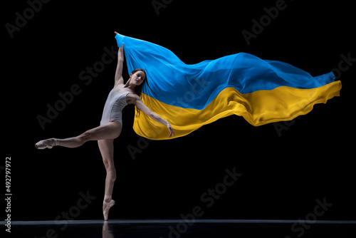 Young graceful classic ballerina dancing with cloth painted in blue and yellow colors of Ukraine flag on dark studio background. Art, peace, freedom and rights