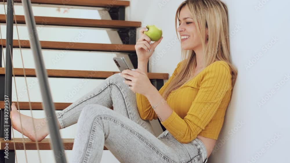 Video of beautiful woman using her smartphone while eating apple sitting on stairs at home.