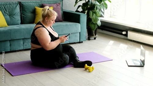 A lazy obese woman is sitting on a sports mat with a phone and does not show desire and activity for the start of a fitness workout. Inactive sedentary person with excess weight photo