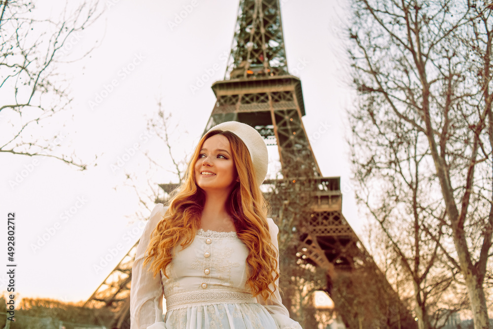 A girl against the backdrop of the Eiffel Tower in Paris in a beret and a dress with curled hair, a romantic journey. Woman laughing and looking away