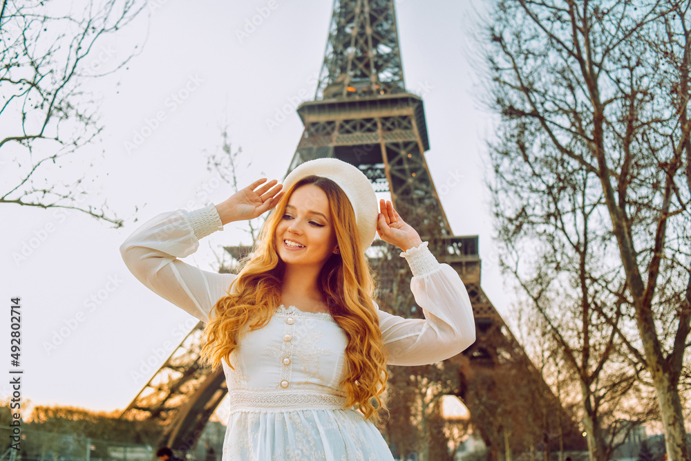 A girl against the backdrop of the Eiffel Tower in Paris in a beret and a dress with curled hair, a romantic journey. Woman laughing and looking away