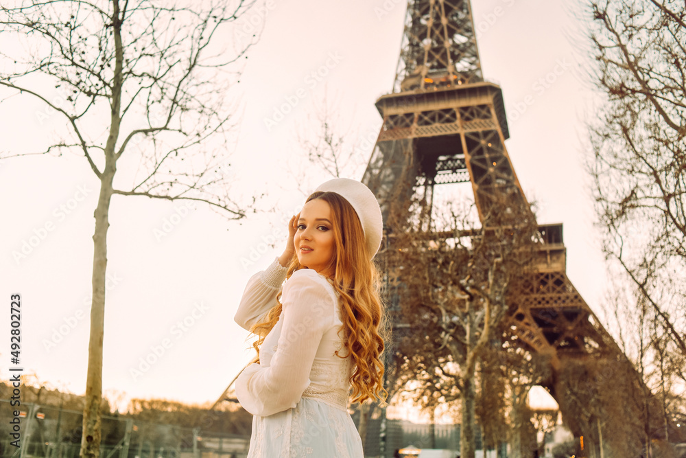 Beautiful blonde in Paris with the Eiffel Tower in the background in a beret and white dress. The woman is laughing and smiling, the girl is on a trip to France