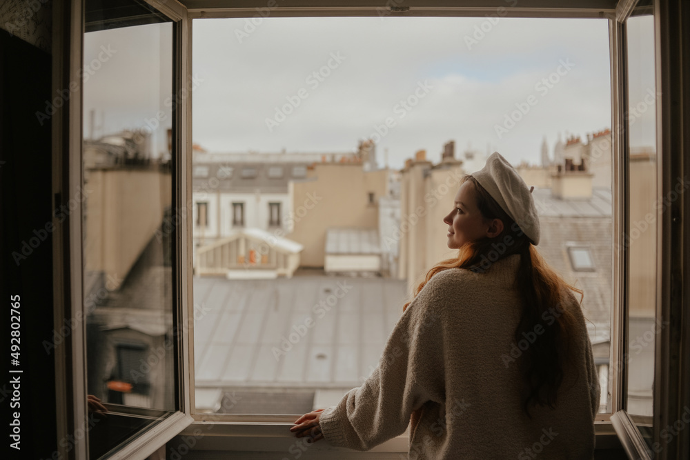 A woman stands near the window against the backdrop of the roofs of Paris. A girl with long hair in a beige suit and beret enjoys the city and the trip